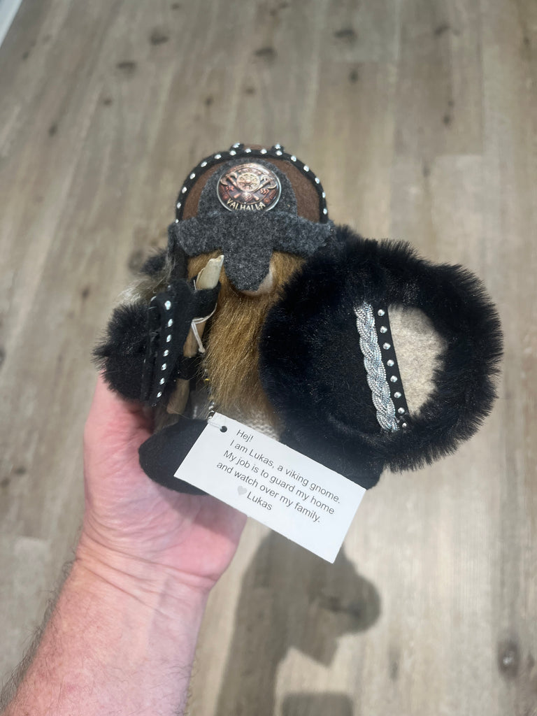 A hand holding a small black and brown plush toy or plush accessory with a gray band, white pom-poms, a circular patch, and a white product tag, against a wood floor background.