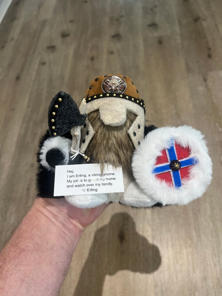 A hand holds a plush Viking gnome toy with a brown fur hat, black and white fur details, a white beard, and a white shield decorated with a red and blue Norwegian flag. A small paper tag with text is attached to the toy.