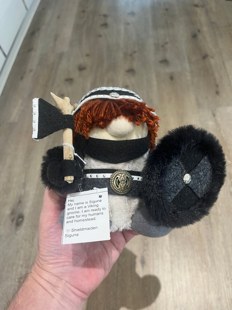 A hand holds a small Viking-themed plush toy with orange hair, a black mask, a shield, and a tag that reads 'Sheld maiden Siguna'. The background shows a wooden floor.