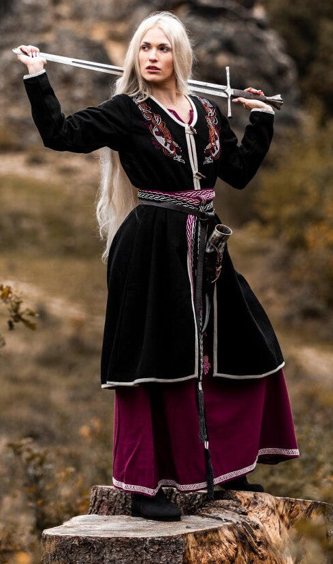 Person in medieval attire holding a sword on a rocky outcrop with trees in the background