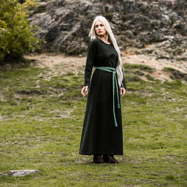 Woman in a long green dress with a green belt standing in a natural landscape with rocks and grass.